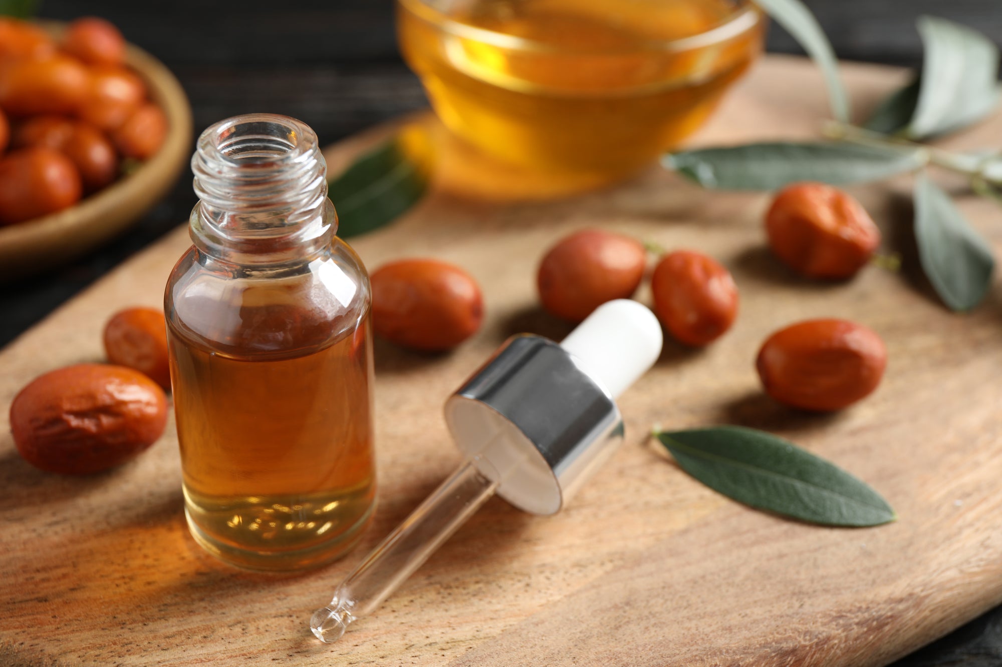 The image showcases a close-up still life composition centered around jojoba oil and its source, the jojoba plant. A small, clear glass bottle with a silver dropper sits prominently in the foreground, filled with a golden-hued liquid – the jojoba oil itself. A single drop hangs delicately from the dropper’s tip. Scattered around the bottle on a light-brown wooden cutting board are several whole, oval-shaped jojoba seeds, displaying a reddish-brown color. A small cluster of these seeds is also visible in a shallow, woven bowl in the upper left corner, suggesting a larger supply. Interspersed among the seeds are a few vibrant green, elongated leaves, likely from the jojoba plant, adding a touch of natural freshness to the scene. Behind the bottle and seeds, a partially visible glass dish contains more of the golden jojoba oil, hinting at the process of extraction or a larger quantity available. The background is a dark, blurred surface, likely a wooden table, which helps to emphasize the warm tones and textures of the foreground elements. The lighting is soft and natural, highlighting the oil’s clarity and the seeds’ smooth surfaces. Overall, the image evokes a sense of natural beauty, wellness, and the origins of a valuable skincare ingredient.